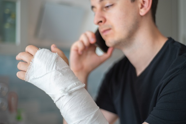 Man with a bandaged arm talking on the phone, possibly at home, with his injury prominently visible in the foreground.